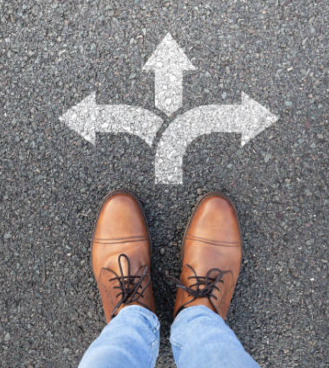 A person wearing brown shoes stands on a paved road facing a painted symbol of three arrows pointing in different directions.